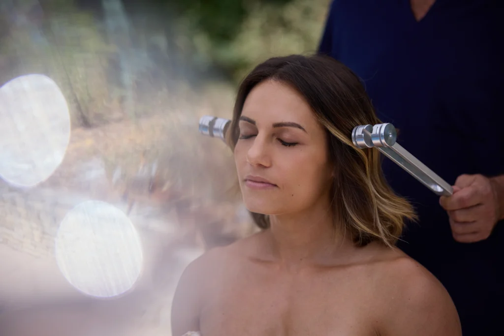 Relaxed client receiving a sound treatment with two tuning forks placed near her ears, enjoying the soothing vibrations.