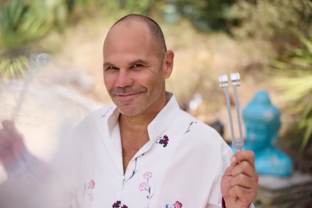 Patrick Gaveau smiling, holding two tuning forks in his hands, used to calm the mind and promote relaxation.